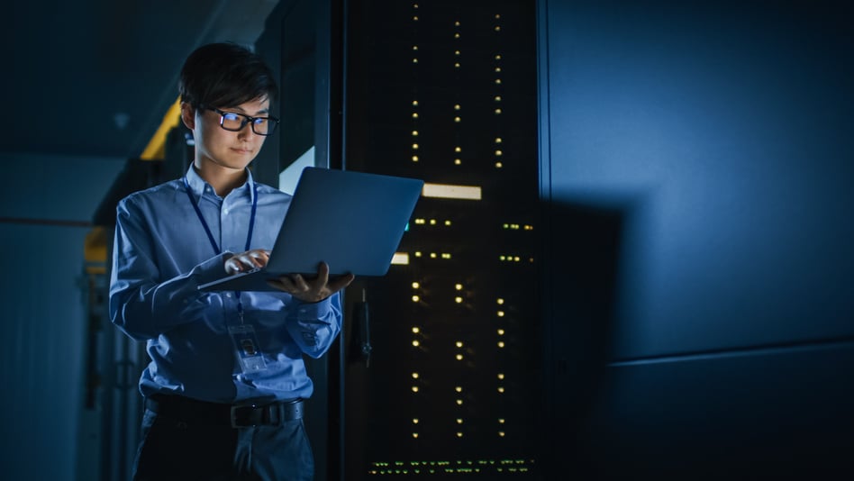 In Dark Data Center: Male IT Specialist Stands Beside the Row of Operational Server Racks, Uses Laptop for Maintenance. Concept for Cloud Computing, Artificial Intelligence, Supercomputer, Cybersecurity. Neon Lights